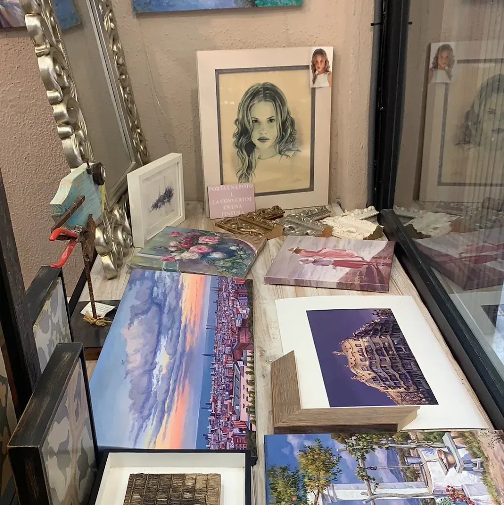 En el escaparate de una tienda de arte se exhibe una muestra de diversas pinturas y retratos, enmarcados y sin enmarcar, que incluyen paisajes y bocetos.