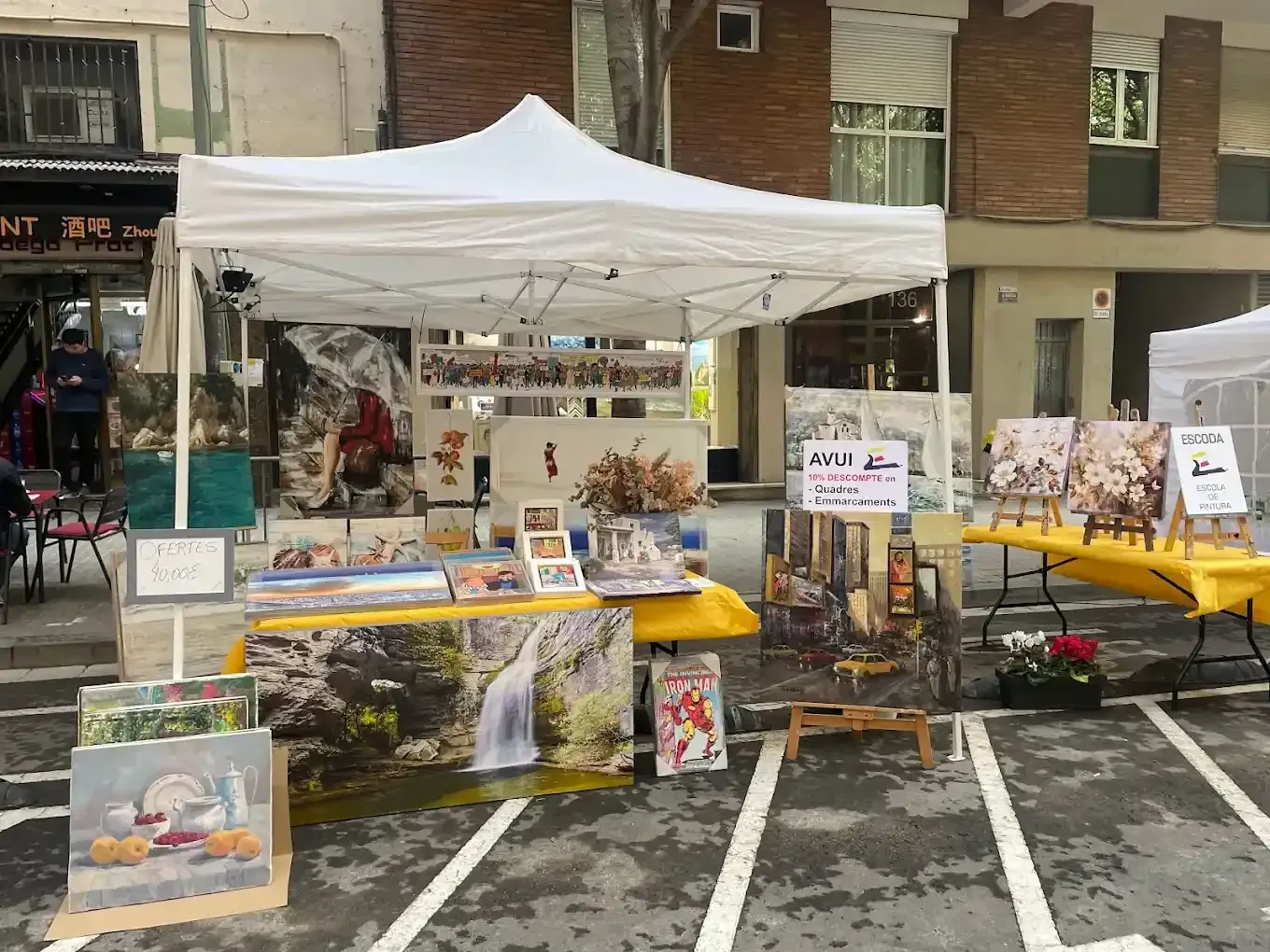 Un puesto de arte al aire libre exhibe pinturas bajo una carpa blanca sobre una superficie pavimentada.