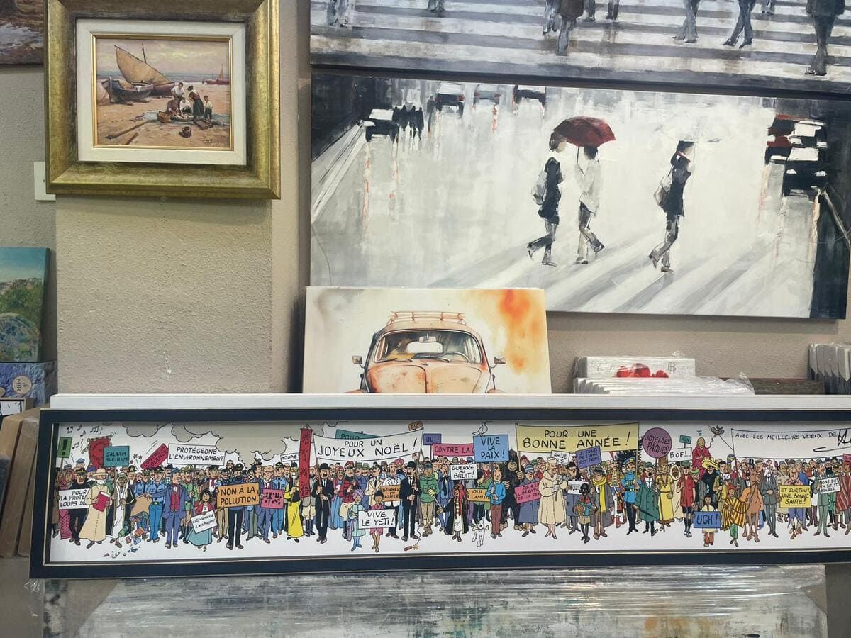 Exposición de arte con cuadros enmarcados que representan una escena de mercado, ciclistas bajo la lluvia y un colorido mural con una multitud.