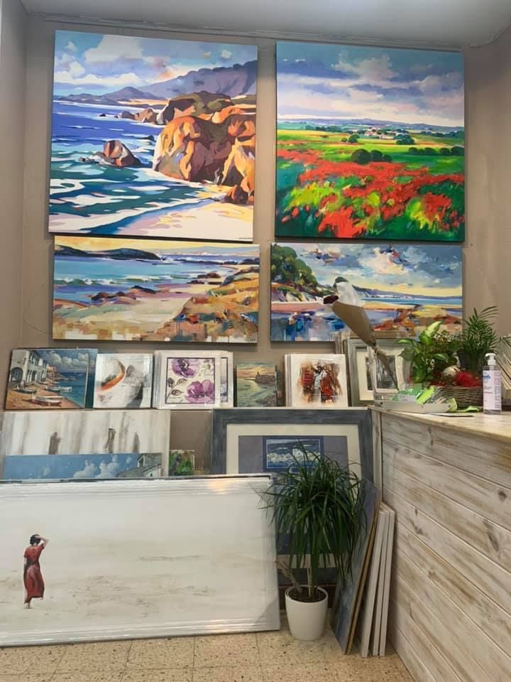 Una galería de arte o tienda que exhibe varios cuadros y grabados de paisajes enmarcados en las paredes y el suelo, con una planta en maceta.