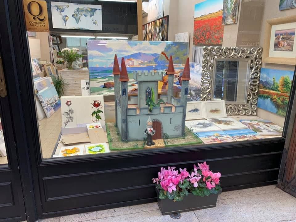 Un castillo de juguete azul se exhibe en el escaparate de una tienda, rodeado de cuadros, un espejo y flores rosas en una maceta.