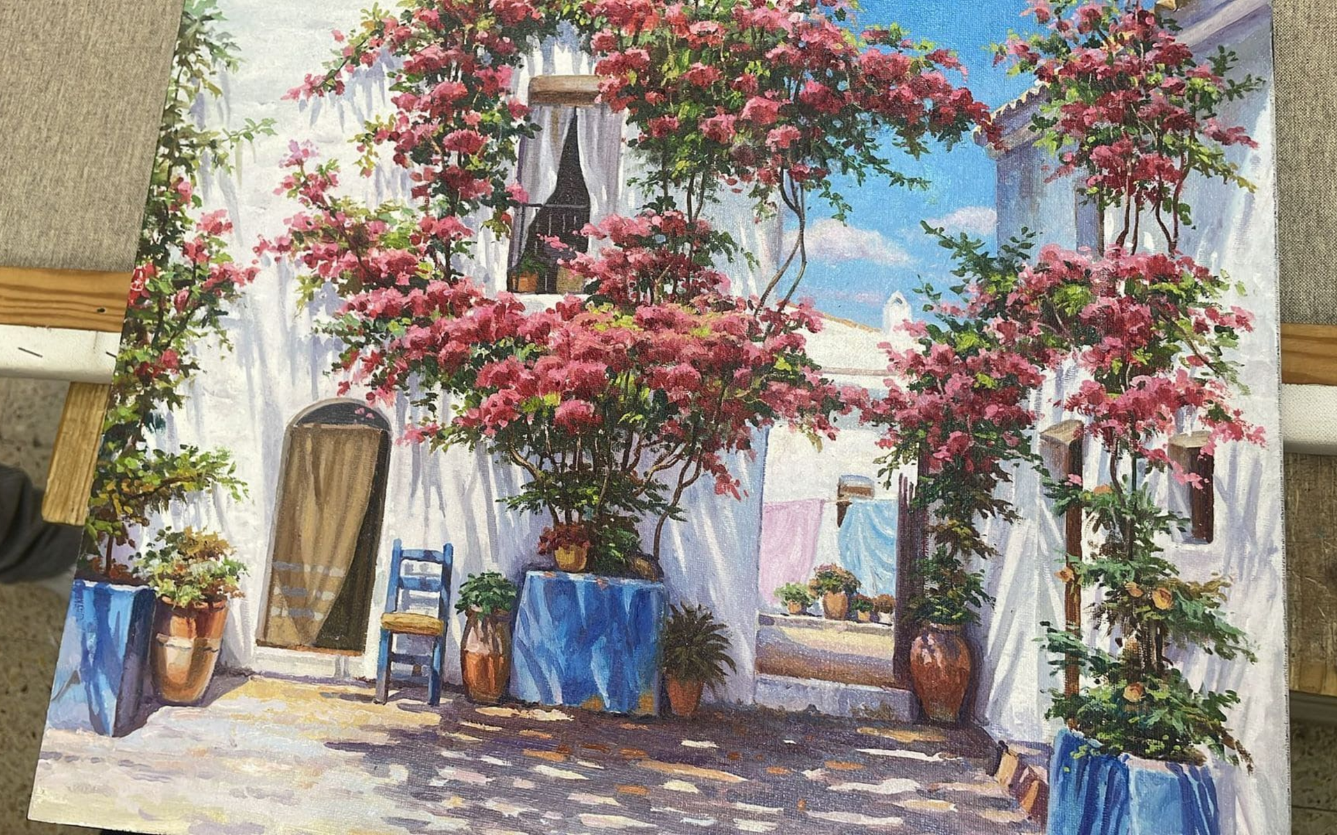 Patio blanco bañado por el sol, con enredaderas de flores rosas, puertas azules y plantas en macetas.