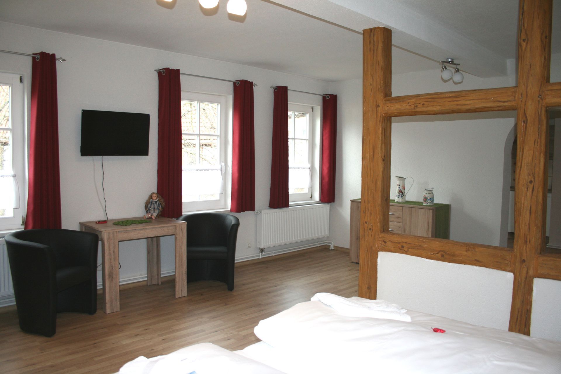 Geräumiges Zimmer mit drei Fenstern, roten Vorhängen, TV, Tisch, Sesseln und Holzbalken.