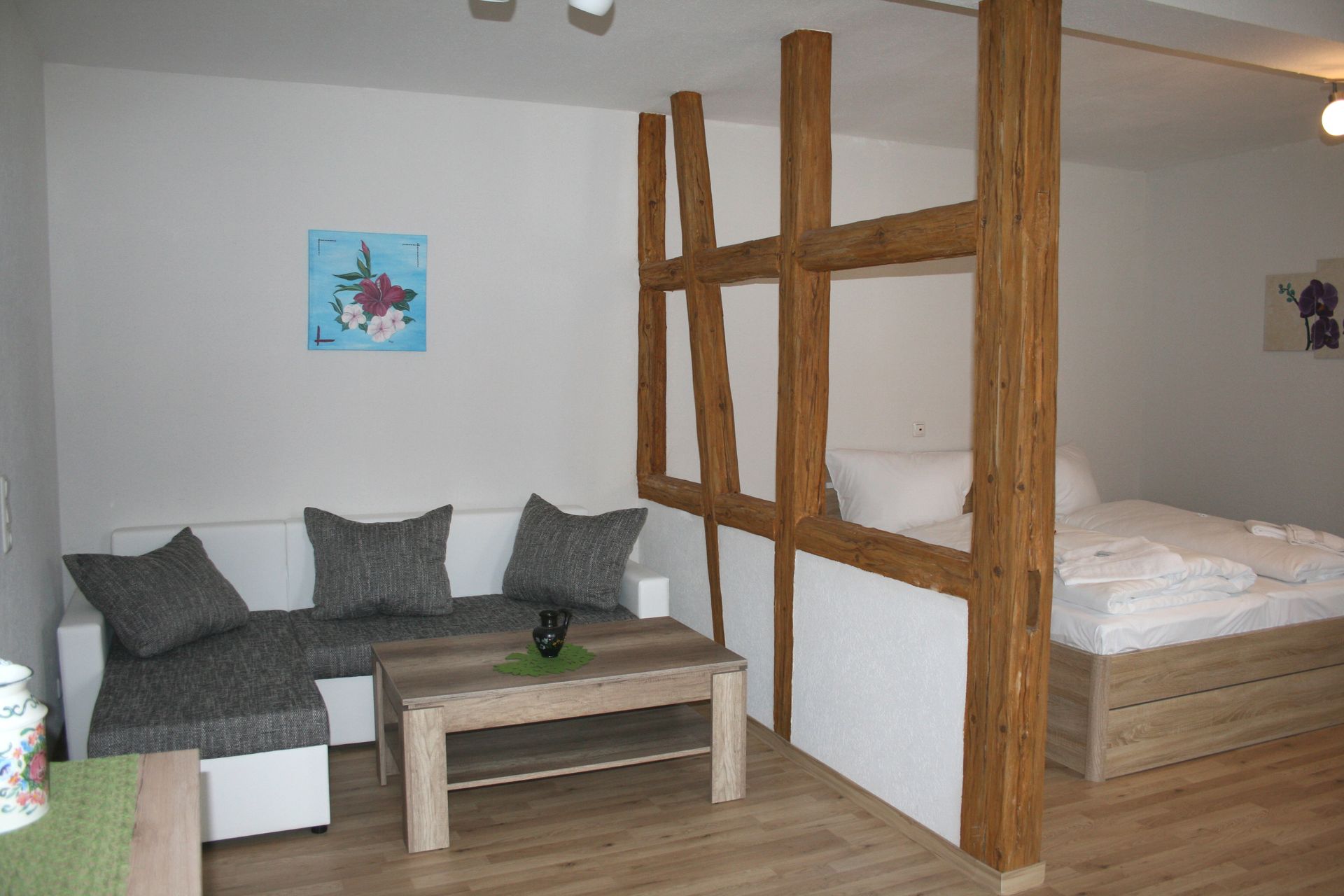 Wohnbereich mit grauem Sofa, Holztisch und Holzbalken als Trennung zum Schlafzimmer.
