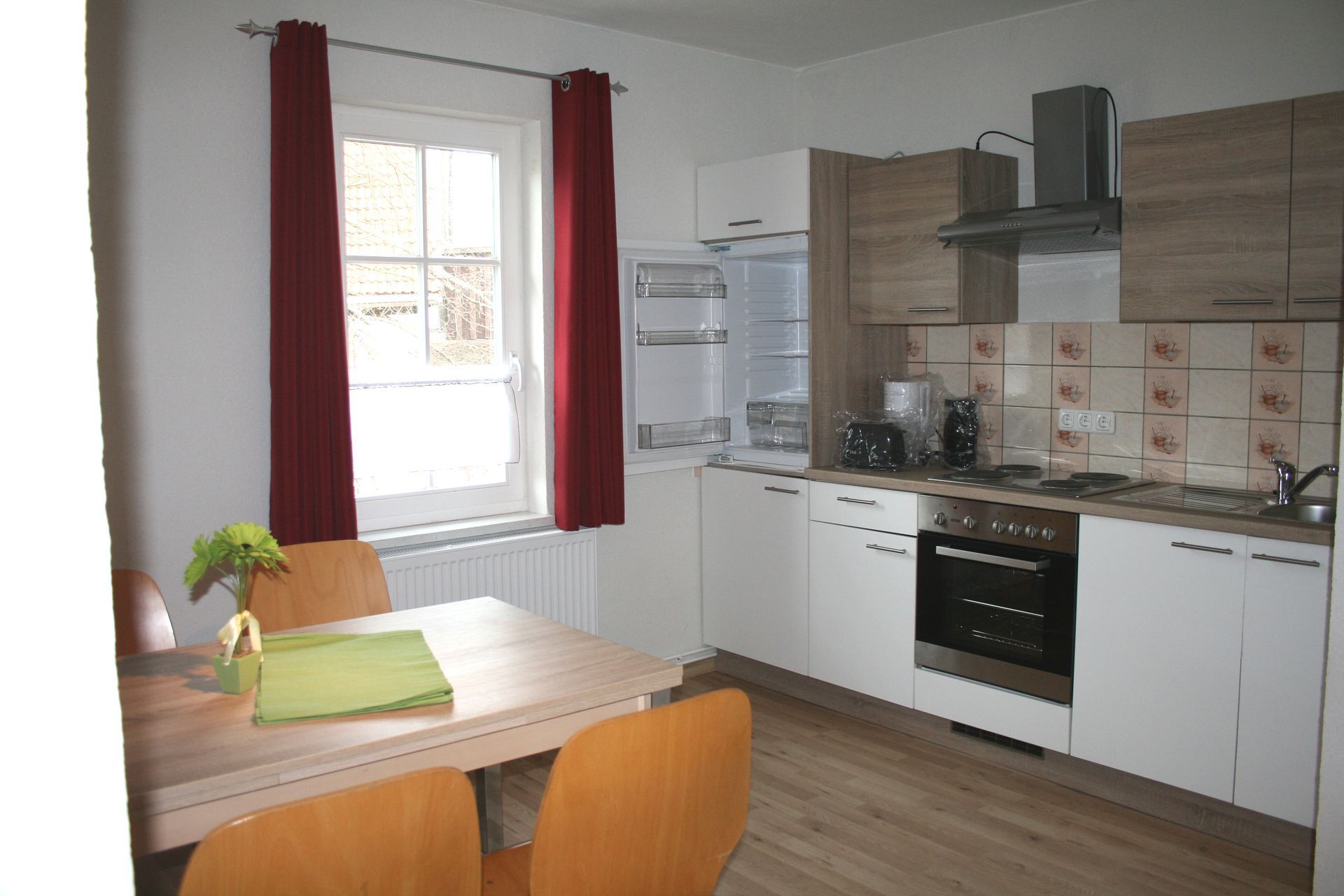 Eine helle Küche mit einem Esstisch aus Holz, modernen Geräten, roten Vorhängen und einem Fenster mit Tageslicht.