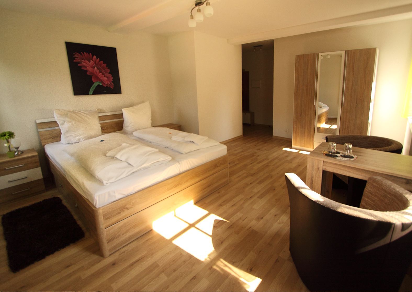 Helles Zimmer mit Doppelbett, Sitzgruppe, Schrank mit Spiegel und rotem Blumenbild an der Wand.