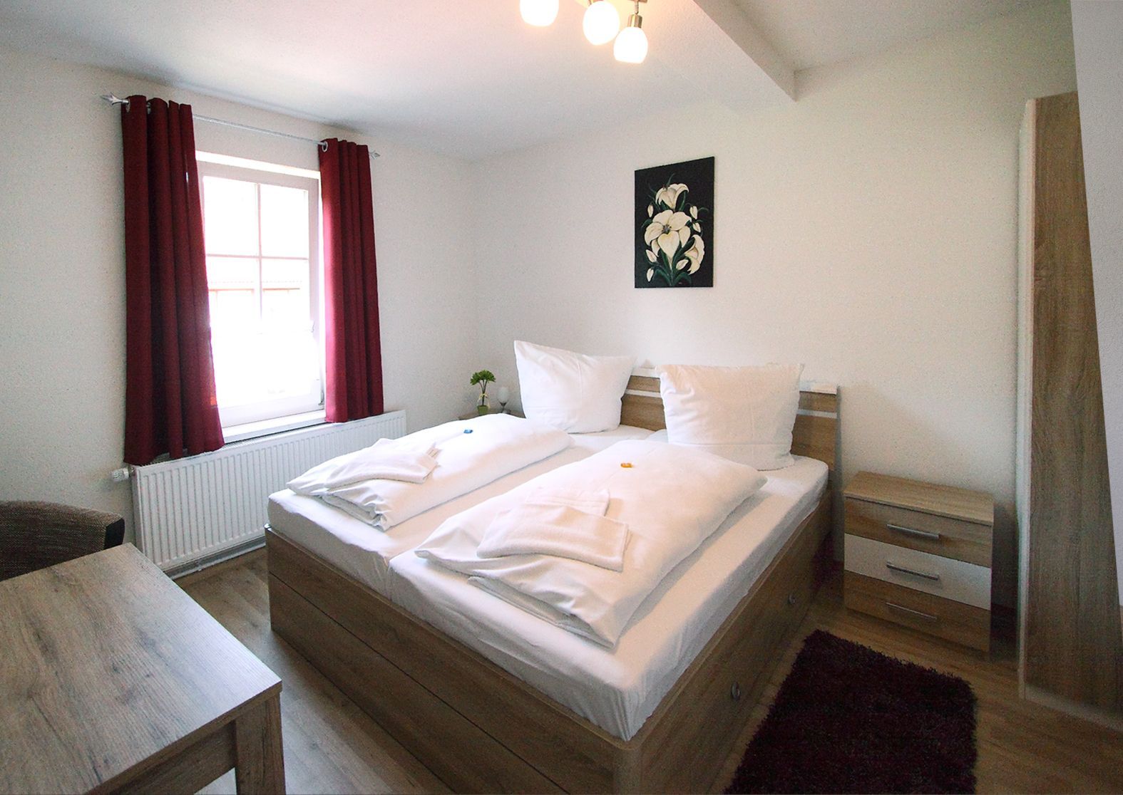 Helles Schlafzimmer mit Doppelbett, Holzmöbeln, weißer Bettwäsche, roten Vorhängen und einem Blumenbild an der Wand.