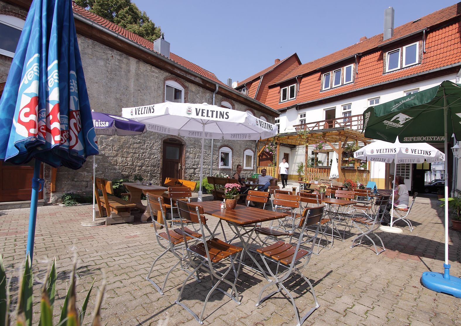 Ein sonniger Außensitzbereich eines Cafés mit Holztischen, Sonnenschirmen und einer rustikalen Gebäudekulisse.