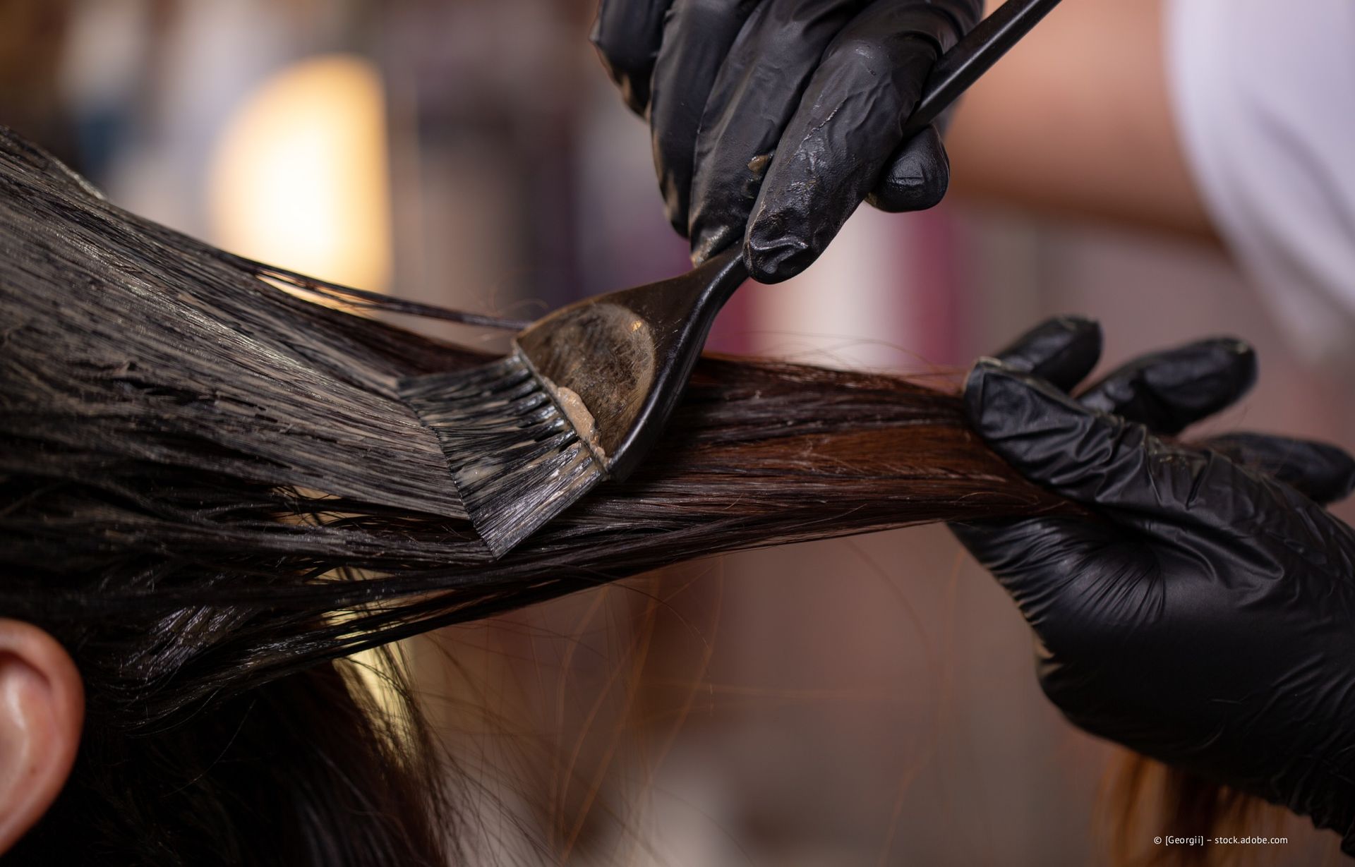 Hände in schwarzen Handschuhen tragen Haarfärbemittel mit einem Pinsel auf.