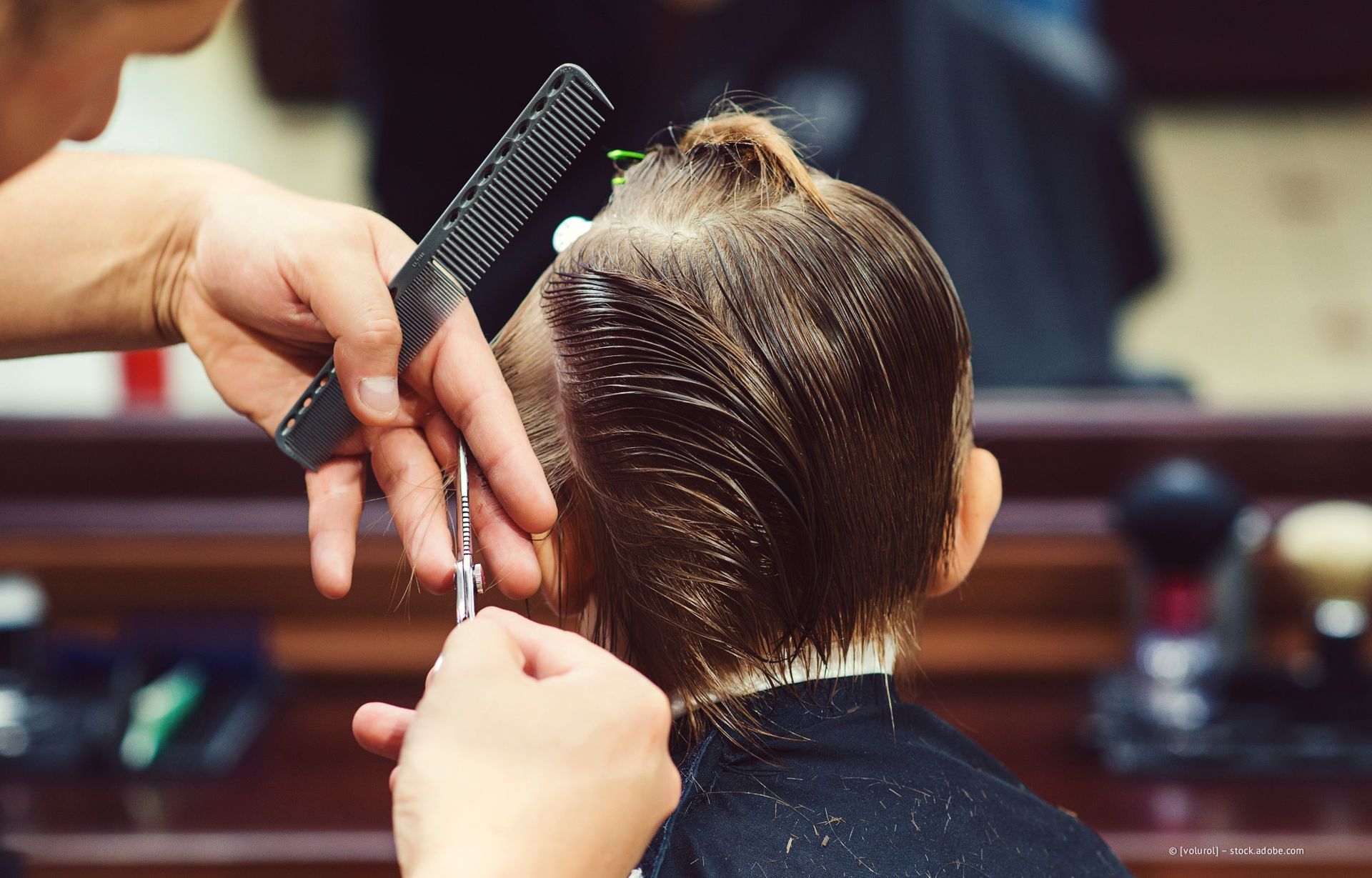 Kind beim Haareschneiden; die Hände des Friseurs halten Schere und Kamm, der Blick ist auf den Hinterkopf gerichtet.