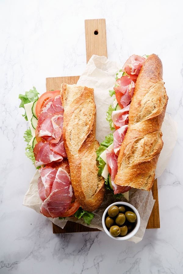 Dos sándwiches con jamón, tomates, lechuga y aceitunas sobre una tabla de cortar de madera.