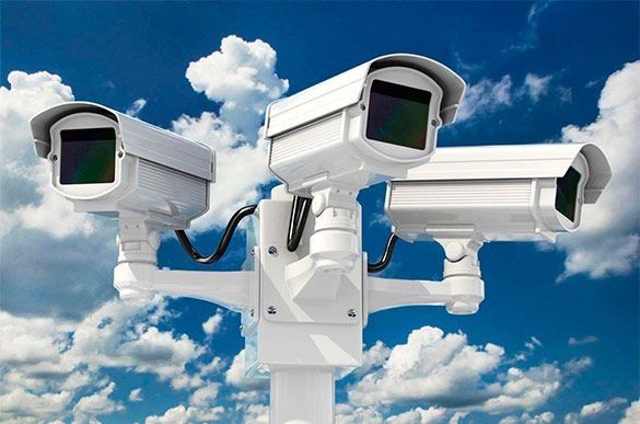 Tres cámaras de seguridad blancas montadas en un poste contra un cielo azul nublado.