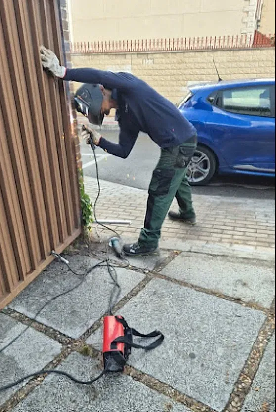 Persona soldando una valla metálica al aire libre, con casco y guantes, con un coche azul y una pared de ladrillos al fondo.