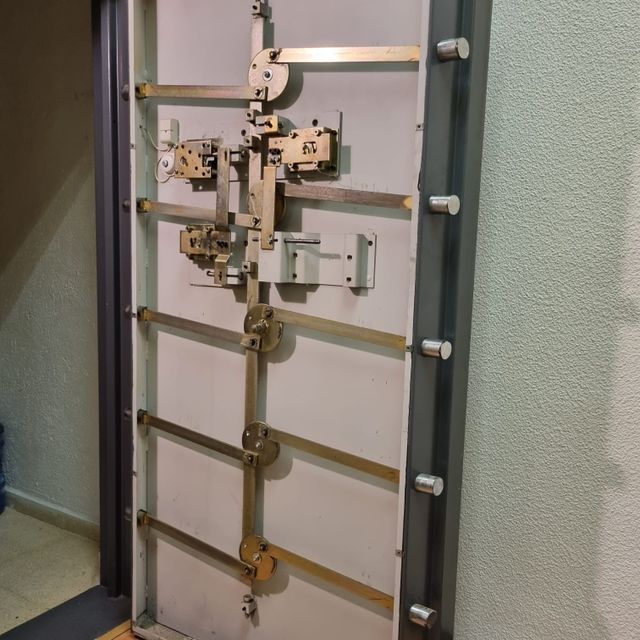 Puerta de bóveda bancaria abierta con mecanismo de bloqueo complejo, gris, dorado y blanco.