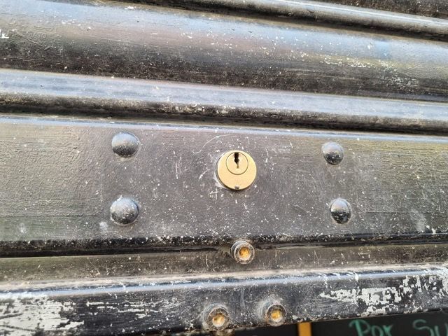 Un primer plano de una persiana metálica negra con cerradura y remaches, probablemente en la fachada de una tienda.