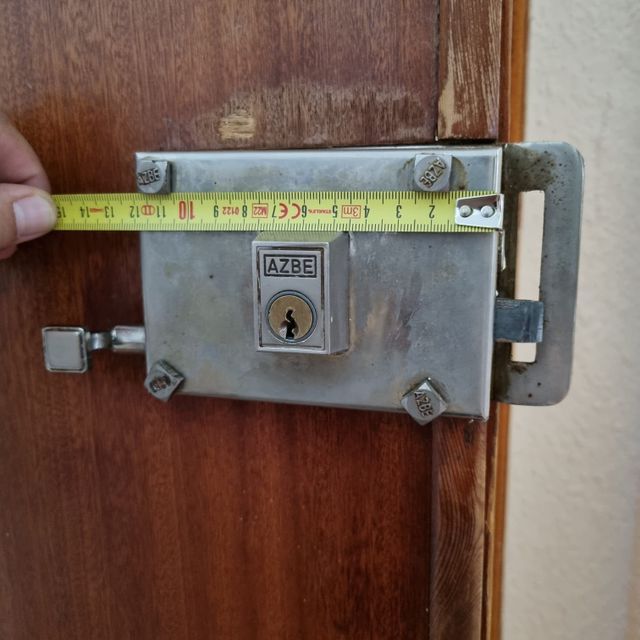 Mano midiendo una cerradura de puerta AZBE plateada en una puerta de madera.