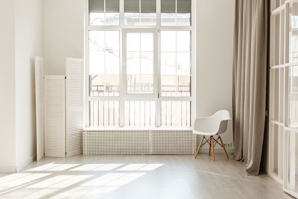 Heller Raum mit großem Fenster, weißen Wänden, Stuhl und Paravent. Sonnenlicht wirft Schatten auf den Holzboden.