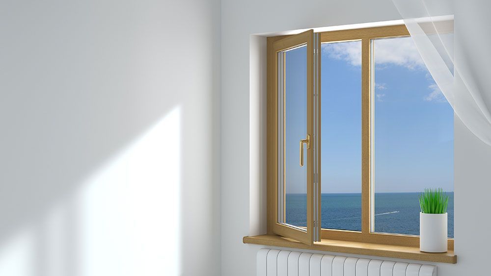 Helles Zimmer mit offenem Fenster mit Blick auf das Meer und den blauen Himmel; auf der Fensterbank steht eine Topfpflanze.