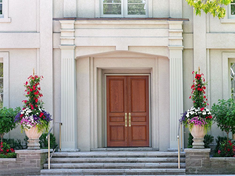 Hölzerne Doppeltüren mit Messinggriffen, eingerahmt von Säulen und Blumentöpfen.