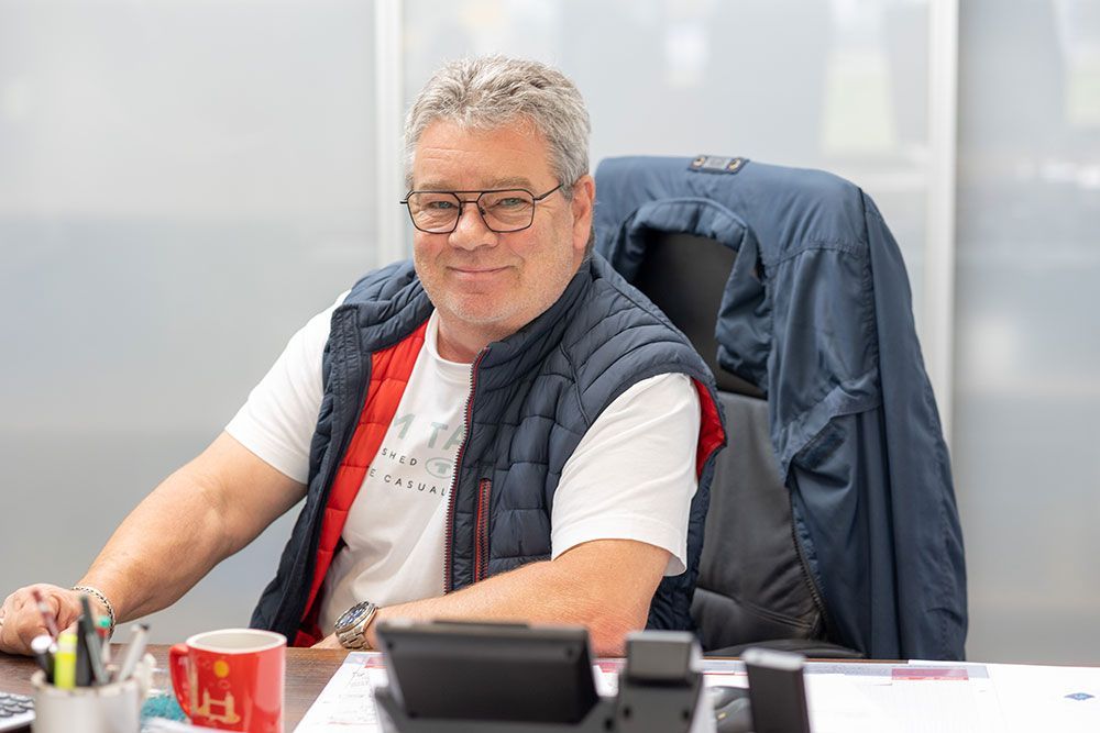 Mann mit Brille am Schreibtisch, trägt Weste, lächelt, mit Tasse und Papieren.