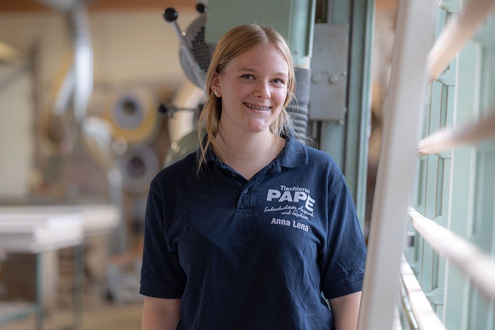 Junge Frau mit Zahnspange lächelt, trägt ein blaues PAPÉ-Shirt und steht neben Industrieanlagen.