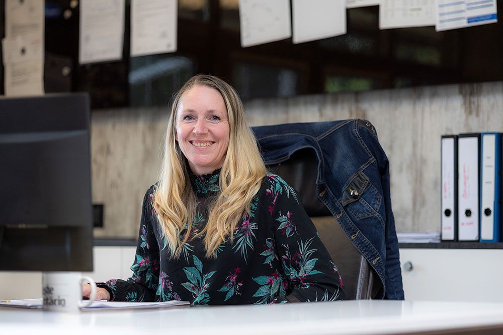 Frau mit blonden Haaren lächelt, während sie an einem Schreibtisch mit einem Computer sitzt. Jeansjacke hängt auf dem Stuhl.