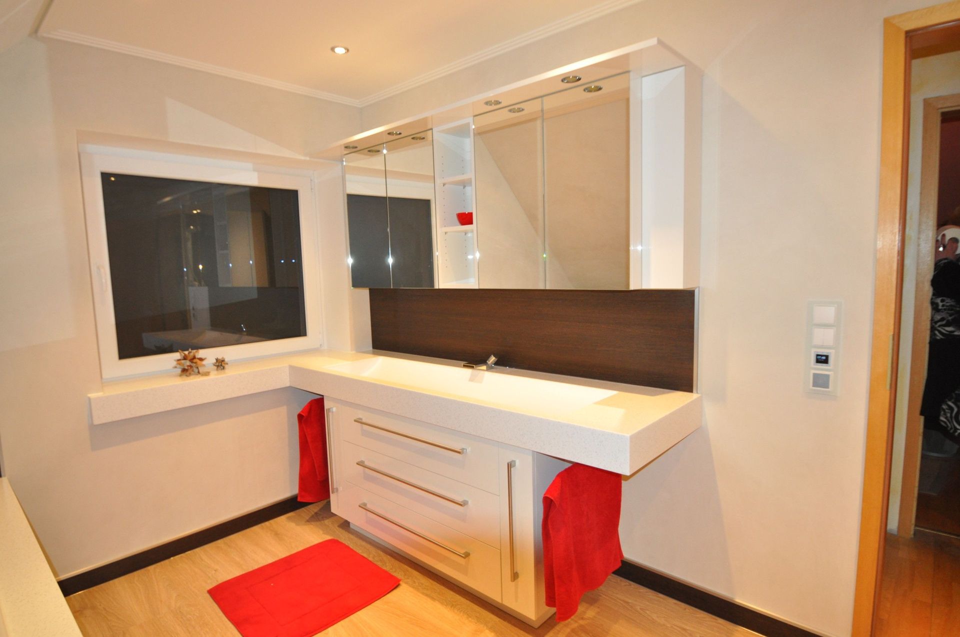 Badezimmer mit weißem Waschtisch, brauner Küchenrückwand, roten Handtüchern und einem Fenster.