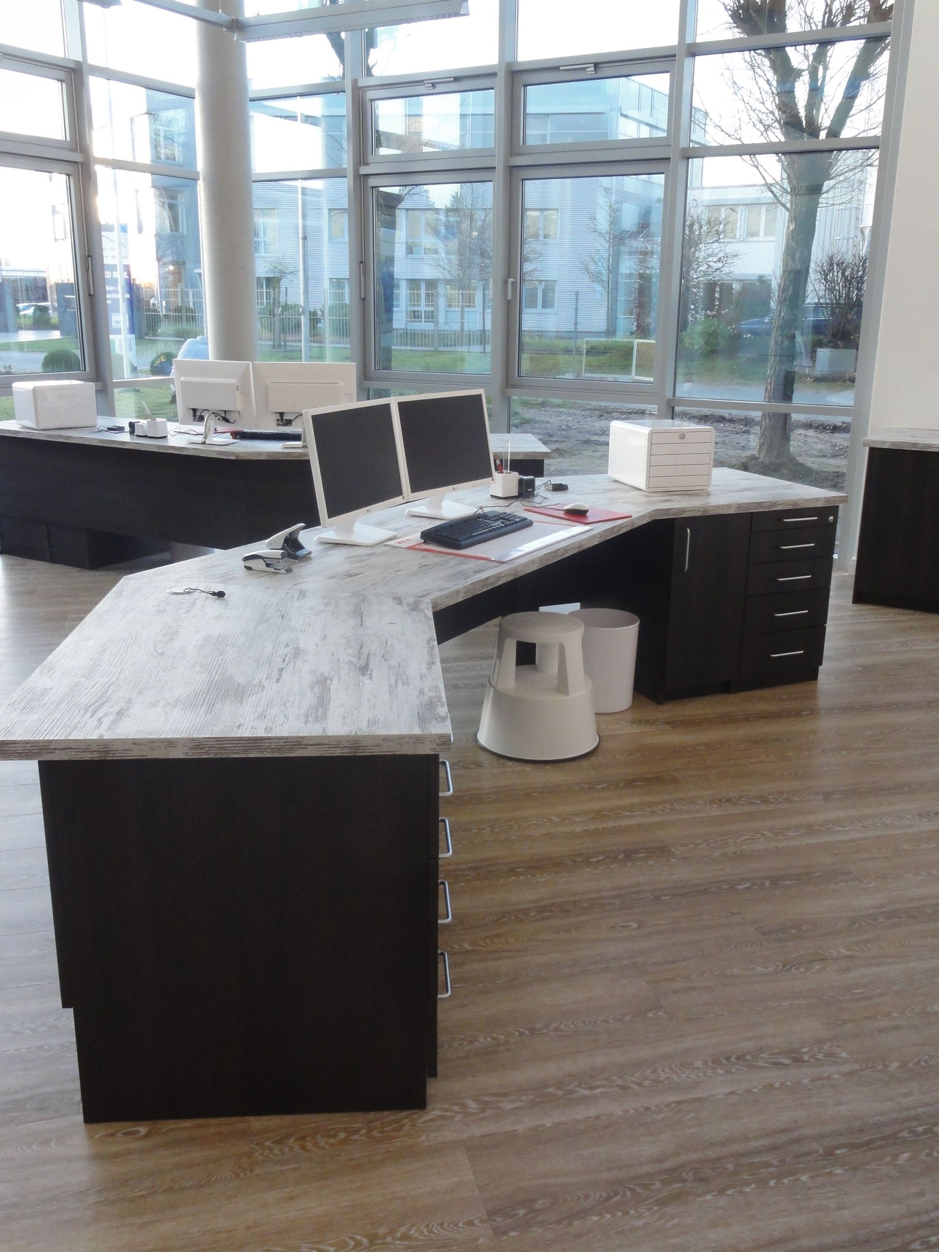 Ein leerer Büroschreibtisch mit Monitoren, Computer und Schubladen in einem modernen Büro mit großen Fenstern.