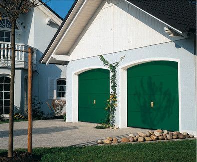 Grüne Garagentore an einem hellblauen Haus, an dessen Wand eine Weinrebe wächst und auf dem Gras Steine liegen.