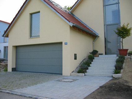 Garagentor und Eingang zu einem modernen beigen Haus, graue Tür, Stufen und Landschaftsgestaltung.