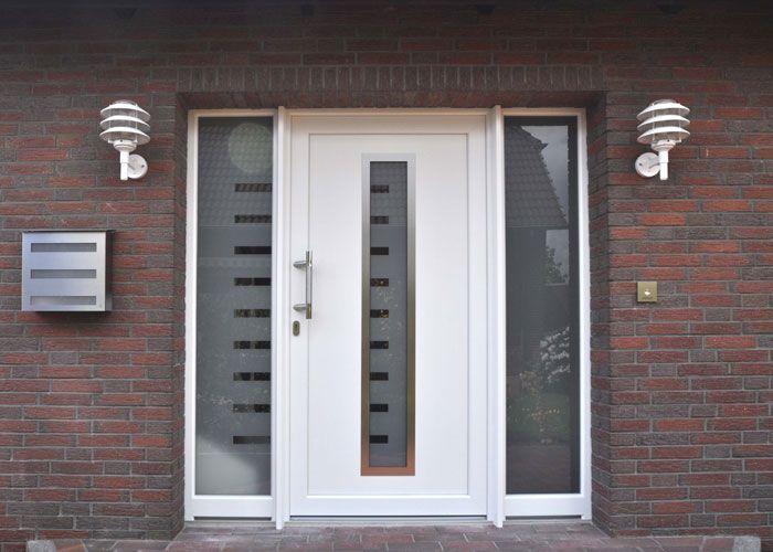 Weiße Haustür mit Glaseinsätzen und Seitenlichtern, flankiert von Wandleuchtern, an einem Backsteingebäude.