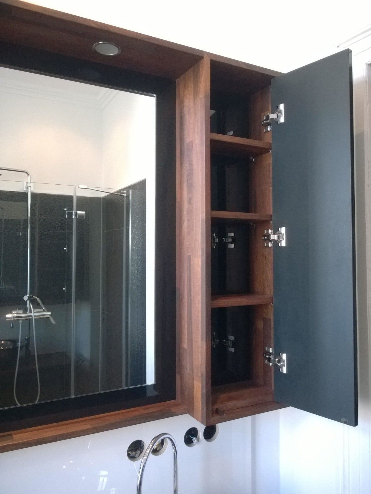 Badezimmerspiegel mit offenem Medizinschrank aus Holz. Spiegel mit schwarzem Rahmen, dunkle Holzverkleidung und Dusche.