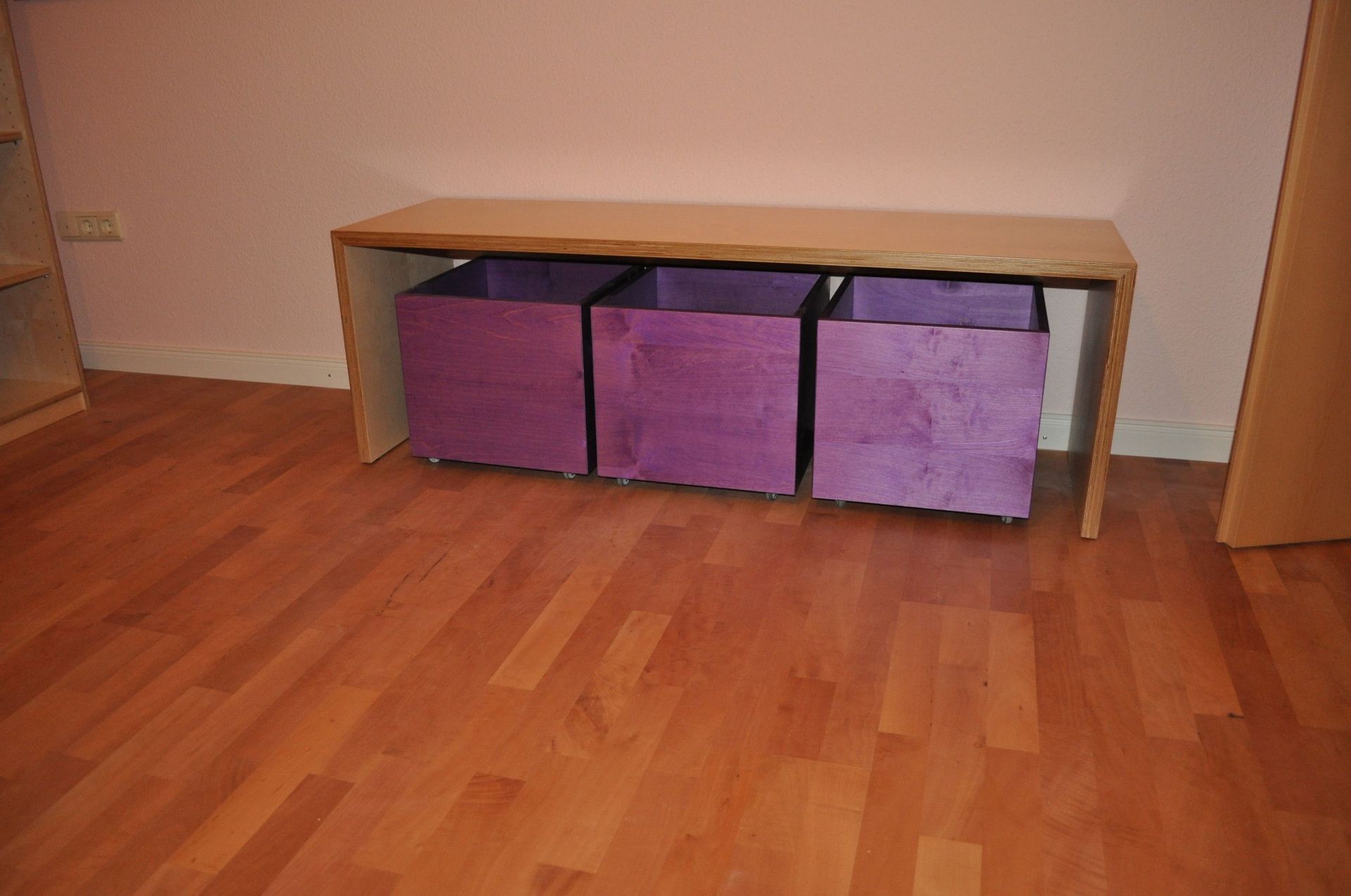 Holzbank mit drei violetten Aufbewahrungswürfeln auf einem Holzboden vor einer rosa Wand.