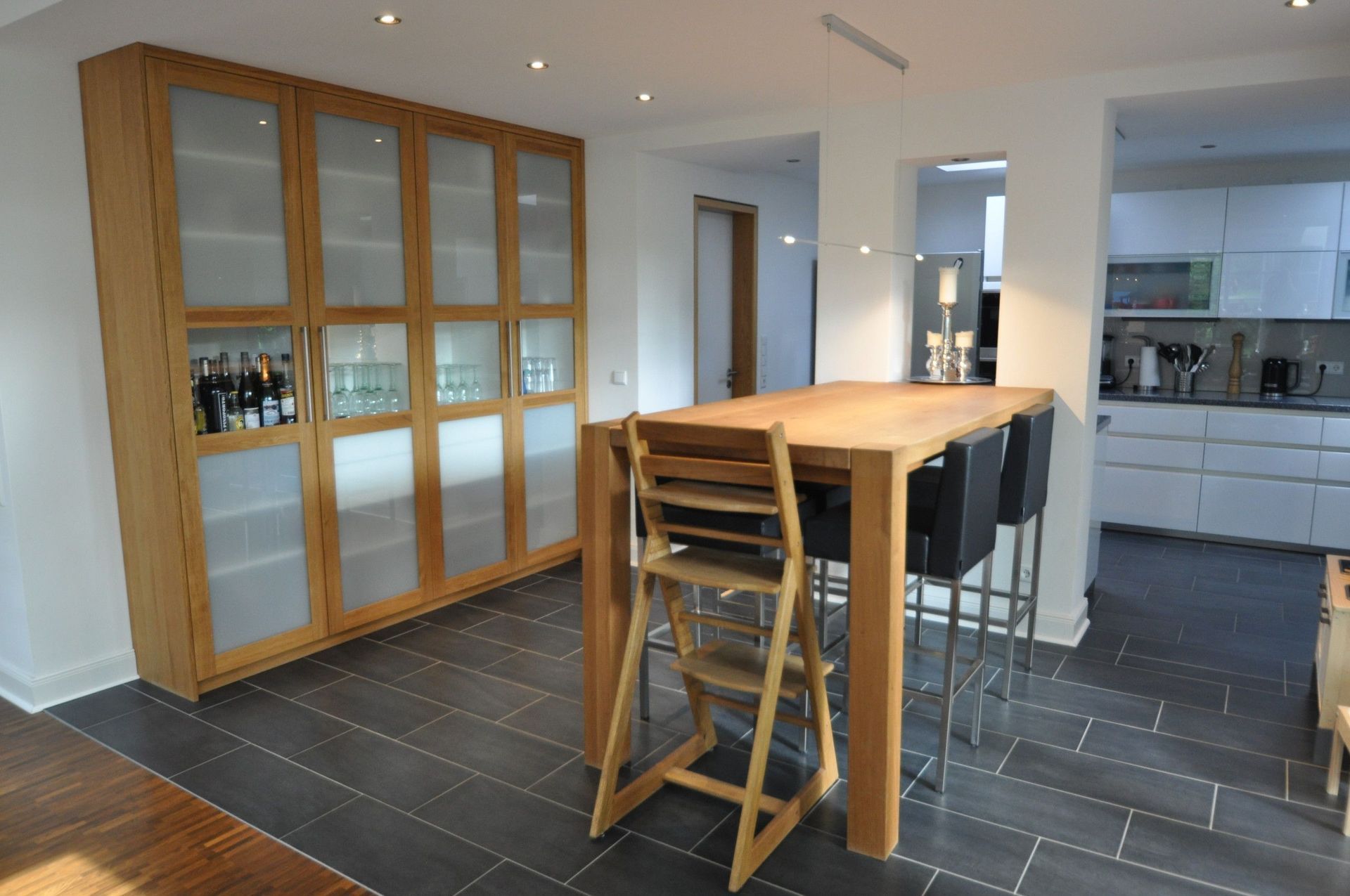 Küche mit Holzschränken, großem Tisch mit Barhockern, Hochstuhl und Blick in eine moderne Küche.