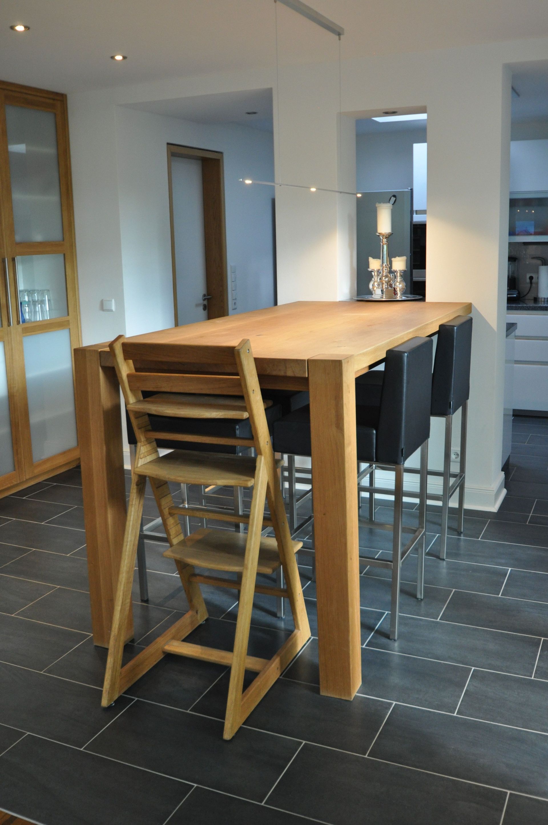 Ein Esstisch mit einem hölzernen Hochstuhl und schwarzen Barhockern; dunkler Fliesenboden.