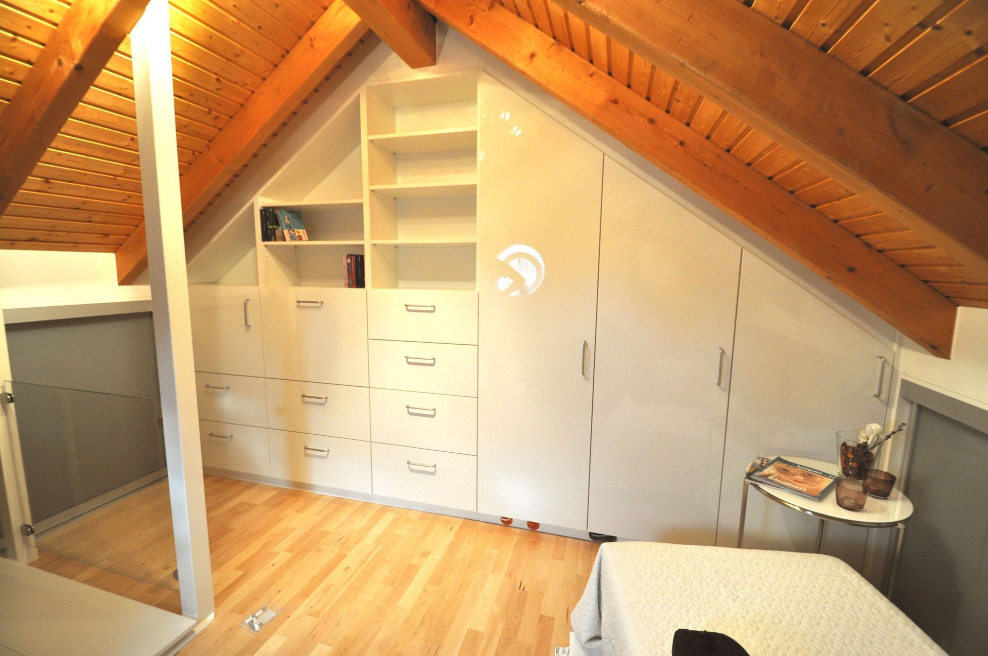 Dachzimmer mit eingebauten weißen Lagerschränken, Holzdecke und -boden. Ein Bett und ein Nachttisch sind sichtbar.