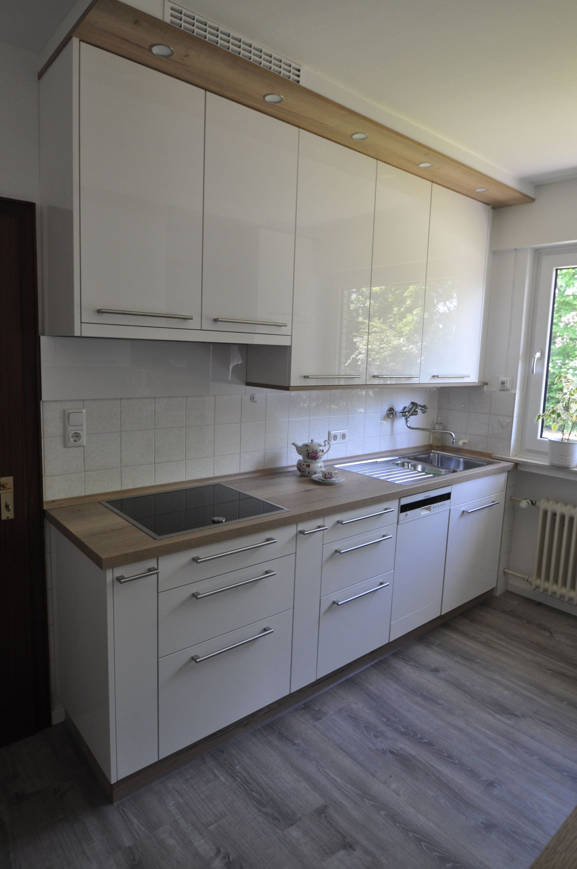 Moderne weiße Küche mit Arbeitsplatten und Schränken aus Holz. Edelstahlbeschläge und ein Fenster mit Blick auf Bäume.