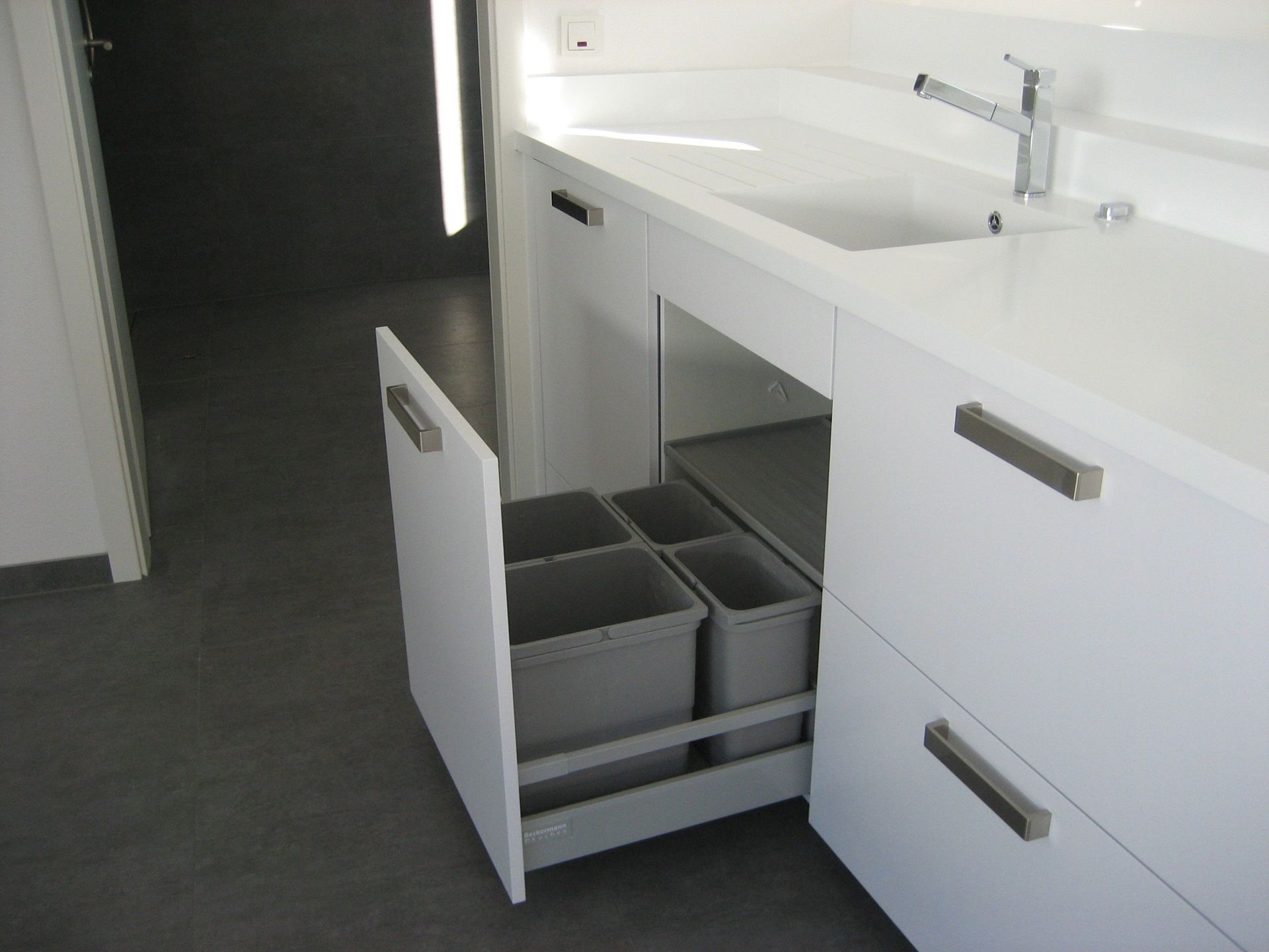 Weißer Küchenschrank mit einer ausziehbaren Schublade mit grauen Mülleimern neben einer weißen Arbeitsplatte und Spüle.