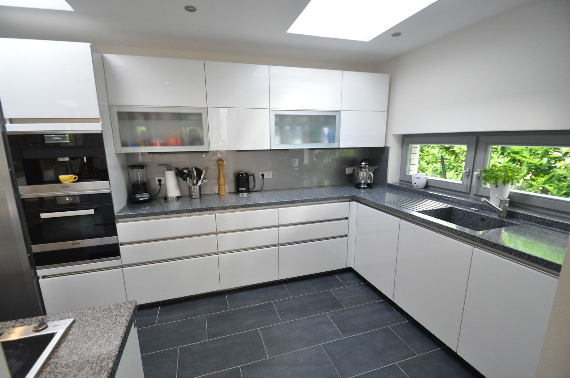 Moderne weiße Küche mit grauen Arbeitsplatten, dunklem Fliesenboden und Fenster mit Blick nach draußen.