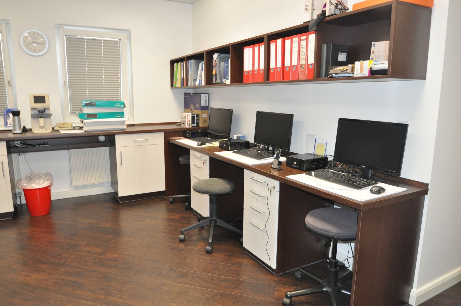 Büro mit Schreibtischen, Computern, Schränken, Regalen mit Ordnern und Büchern, einem Fenster und dunklem Holzboden.