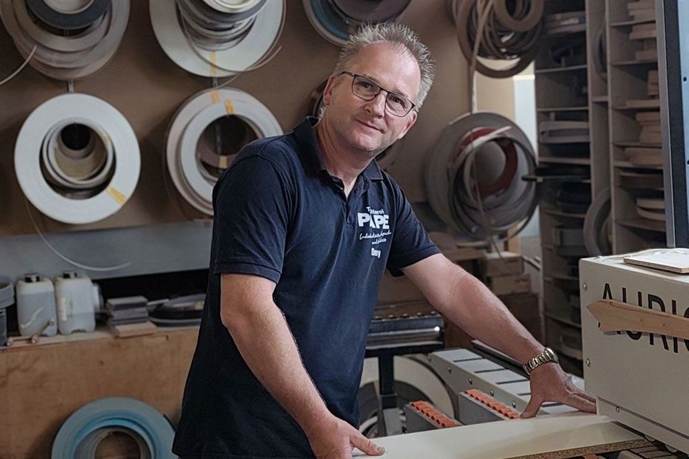Mann mit Brille und blauem Hemd in der Werkstatt arbeitet an einer Maschine, umgeben von Kantenbandrollen.