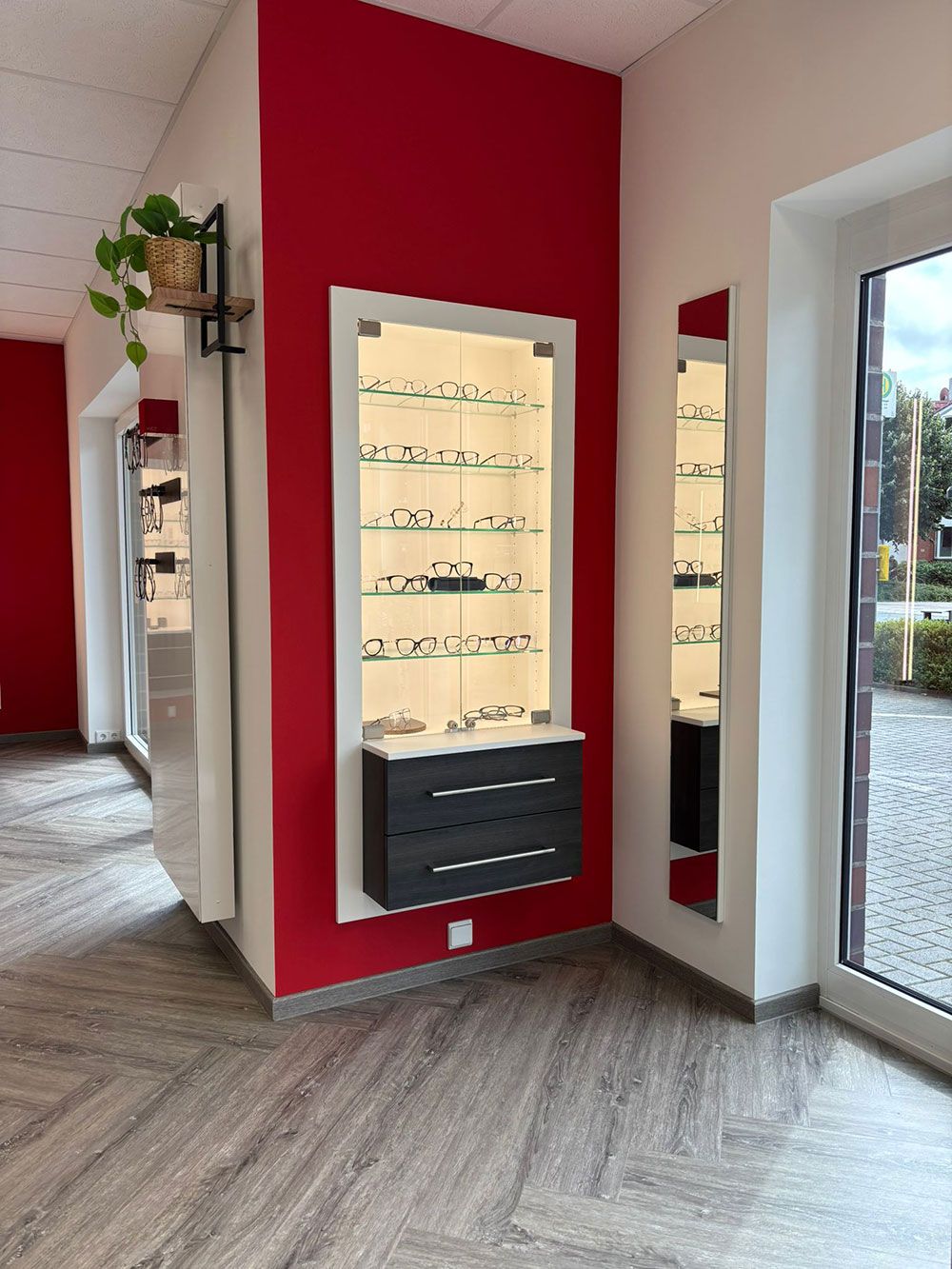 Inneneinrichtung eines Optikergeschäfts mit roter Wand und Glasvitrinen, in denen Brillen präsentiert werden.
