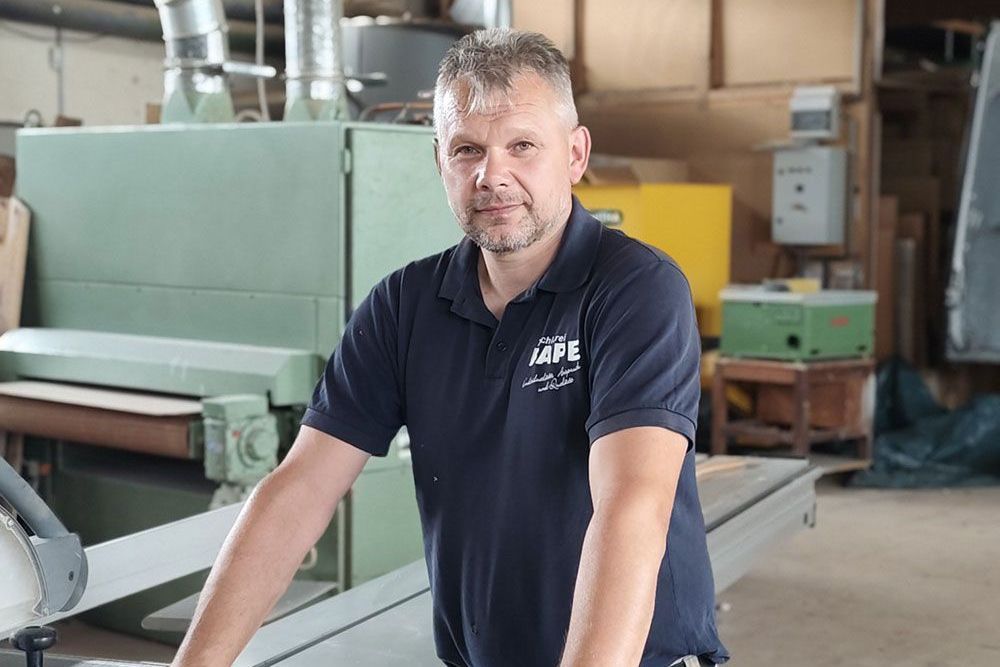Mann in einer Werkstatt, trägt ein marineblaues Poloshirt. Er steht neben Maschinen und blickt in die Kamera.