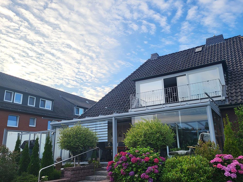 Ein Haus mit glasüberdachter Terrasse, Balkon und Garten unter einem bewölkten blauen Himmel.