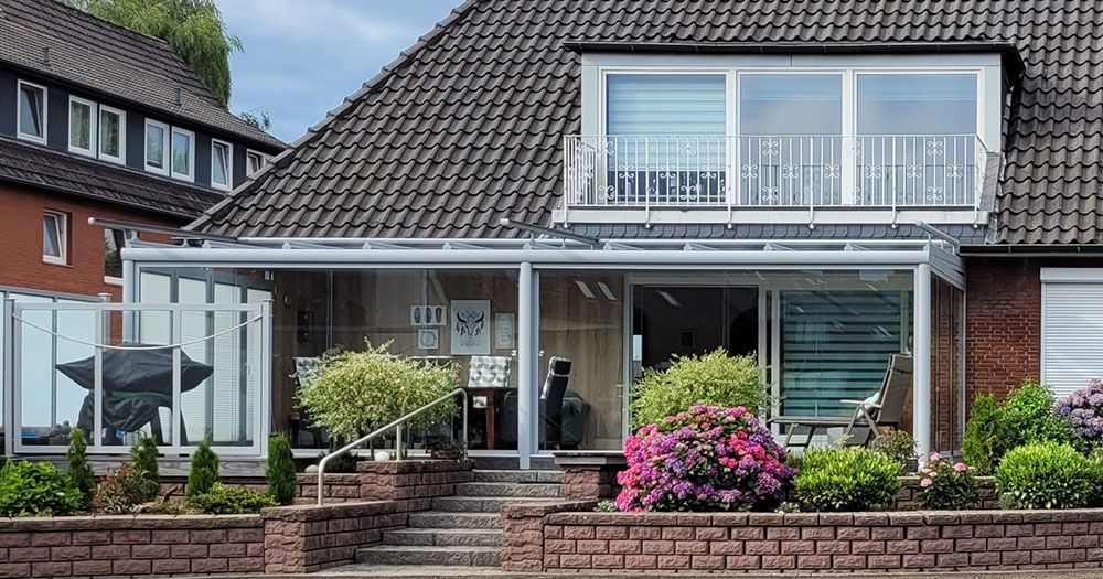 Ein Haus mit einer glasüberdachten Terrasse, Blumenbeeten und einem Balkon im zweiten Stock.