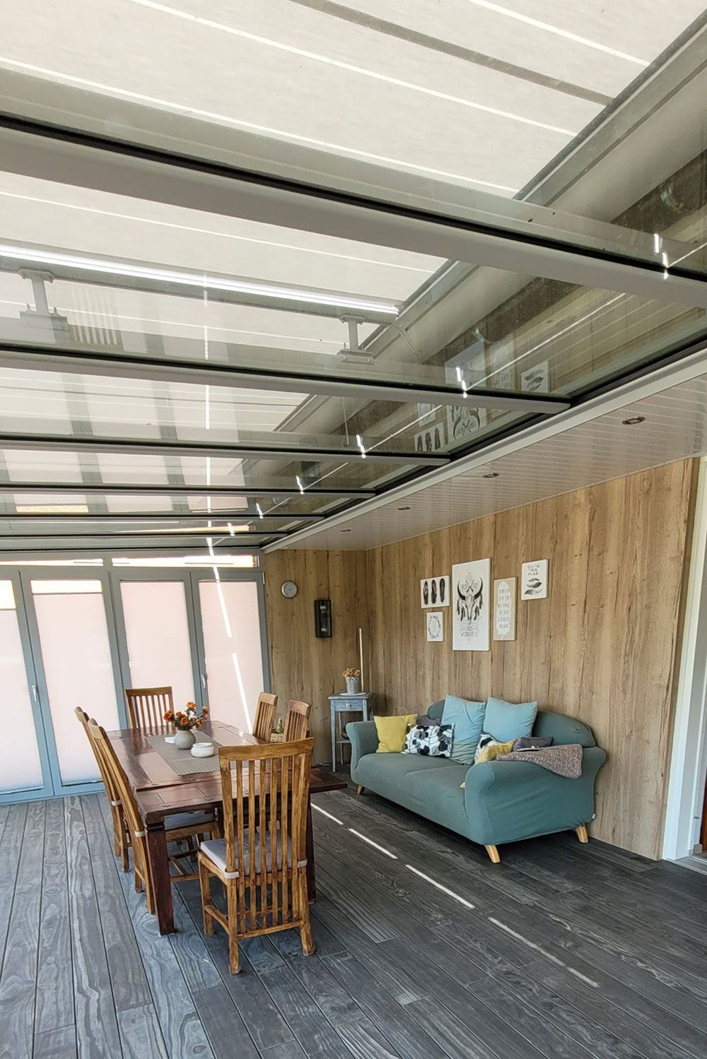 Wintergarten mit Holzpaneelwand, Esstisch, blauem Sofa. Glasdecke und Holzboden.