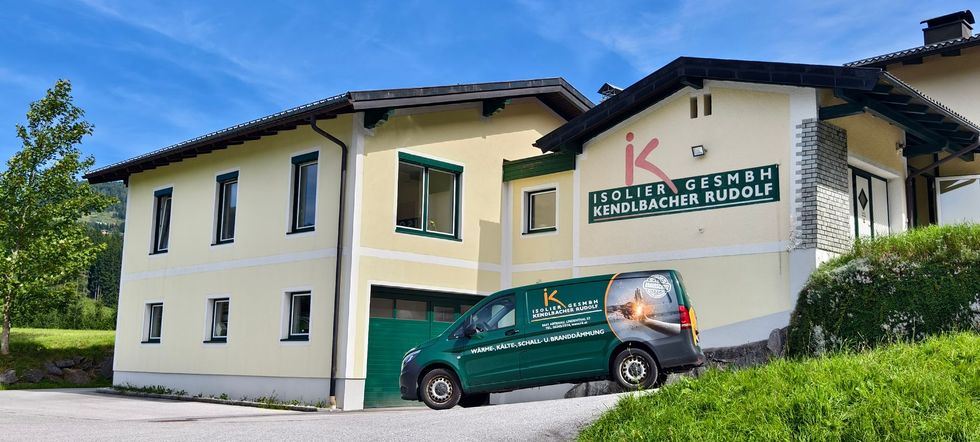 Ein cremefarbenes Gebäude mit grünen Zierelementen und ein grüner Lieferwagen, der davor an einem sonnigen Hang parkt.