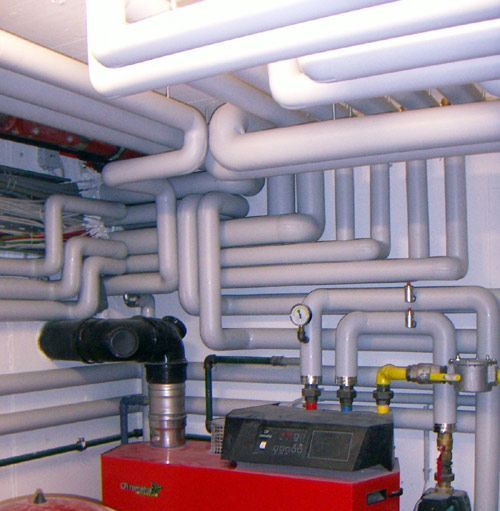 Heizraum mit roter Heizeinheit und dicht verlegten, weißen, isolierten Rohren an Wänden und Decke