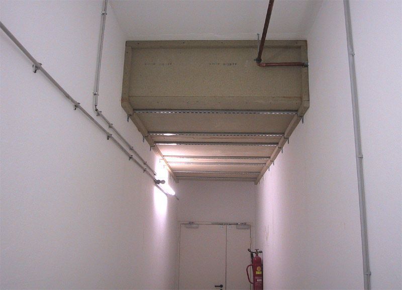 Ein schmaler, weißer Flur mit großen, beigefarbenen Lüftungsrohren, die von der Decke hängen, und einem Feuerlöscher am Ende.