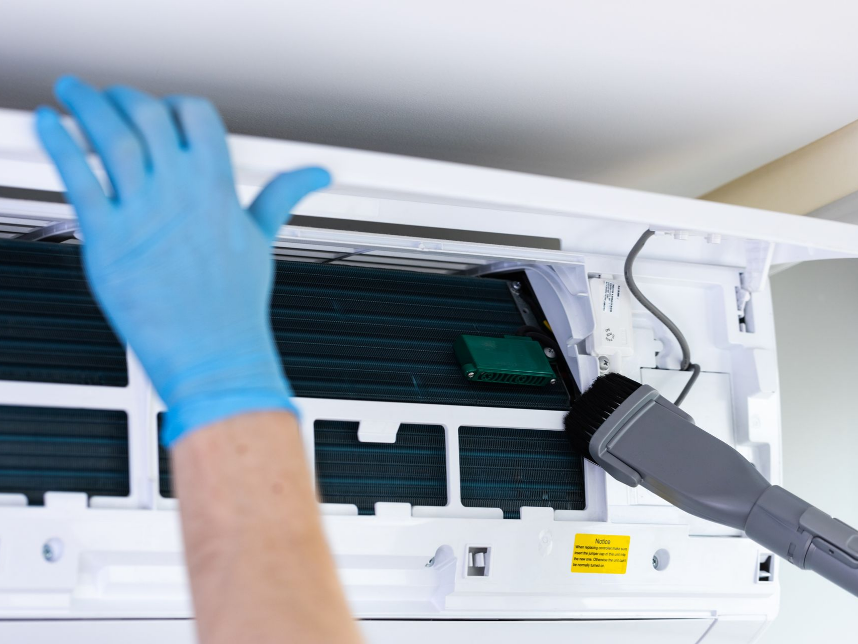 Une personne portant un gant bleu nettoie un climatiseur avec un aspirateur.